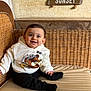 baby, child, smile, hoodie, bear_graphic, wicker_sofa, cushion, indoor, signs, wooden_signs, text, cozy, happy, seated, cute, portrait, infant, clothing, face, person