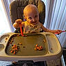 baby, child, high_chair, mealtime, smiling, happy, indoor, curtains, tray, food, vegetables, peas, carrots, spoon, eating, infant, portrait, sitting, home, playful