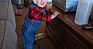 Wesley is registered to the contest to win money with this photo: toddler, child, helmet, smiling, indoors, home, overalls, plaid_shirt, red_shirt, denim, standing, portrait, coffee_table, cardboard_boxes, spray_bottle, vacuum_cleaner, couch, carpet, toys, happy