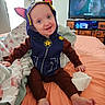Wesley is registered to the contest to win money with this photo: baby, child, infant, smiling, portrait, superhero_costume, hood, hat, blue_clothing, brown_clothing, sitting, bed, blanket, pillow, bedroom, television, furniture, indoor, barefoot, happy