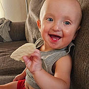Wesley is registered to the contest to win money with this photo: baby, child, smiling, happy, portrait, indoor, couch, sitting, bread, food, hand, blue_eyes, gray_shirt, red_shorts, bald, laughing, infant, home, closeup, casual