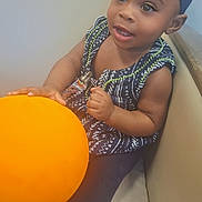 Kalaya is registered to the contest to win money with this photo: toddler, child, baby, blue_headband, orange_ball, sitting, couch, curious, happy, indoor, clothing, patterned_dress, legs, hand, face, smile, person, young_child, cute, portrait