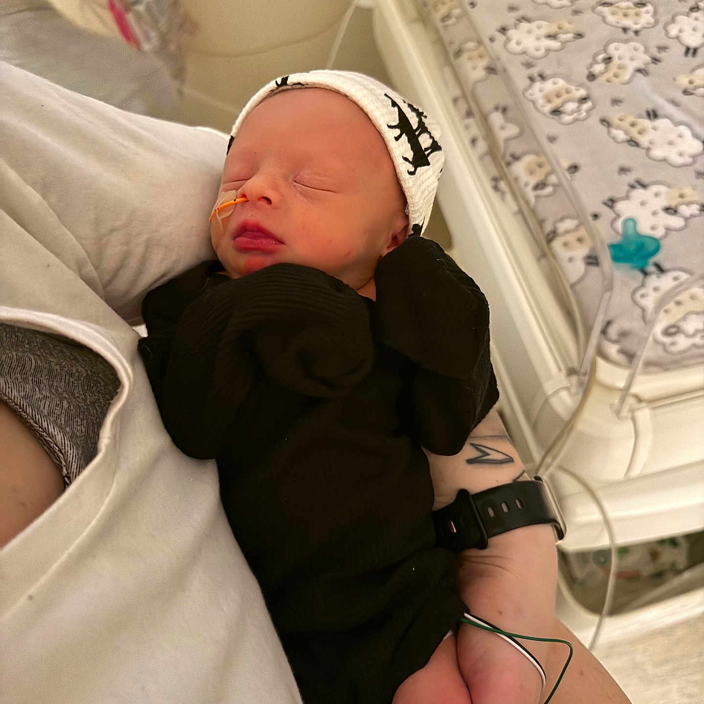 Creed is registered to the contest to win money with this photo: arm, baby, bedroom, black_clothing, blanket, care, cradle, crib, hand, hat, indoor, infant, medical_tube, newborn, pacifier, pattern, person, sleeping, tattoo, watch