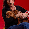 Alba a rejoint le concours — aidez-le/la à gagner de superbes lots ! woman, dog, glasses, red_background, yellow_cushion, casual_clothing, black_shirt, blue_jeans, bracelet, sitting, portrait, pet, expression, indoor, medium_hair, brown_dog, holding, seated, studio_photo, surprised_face