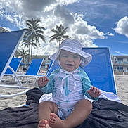 Renzo joined the competition — help win amazing prizes! baby, child, beach, sun_hat, swimsuit, towel, beach_chair, sand, palm_trees, sky, clouds, smiling, happy, outdoor, vacation, summer, sunny, person, foot, hand