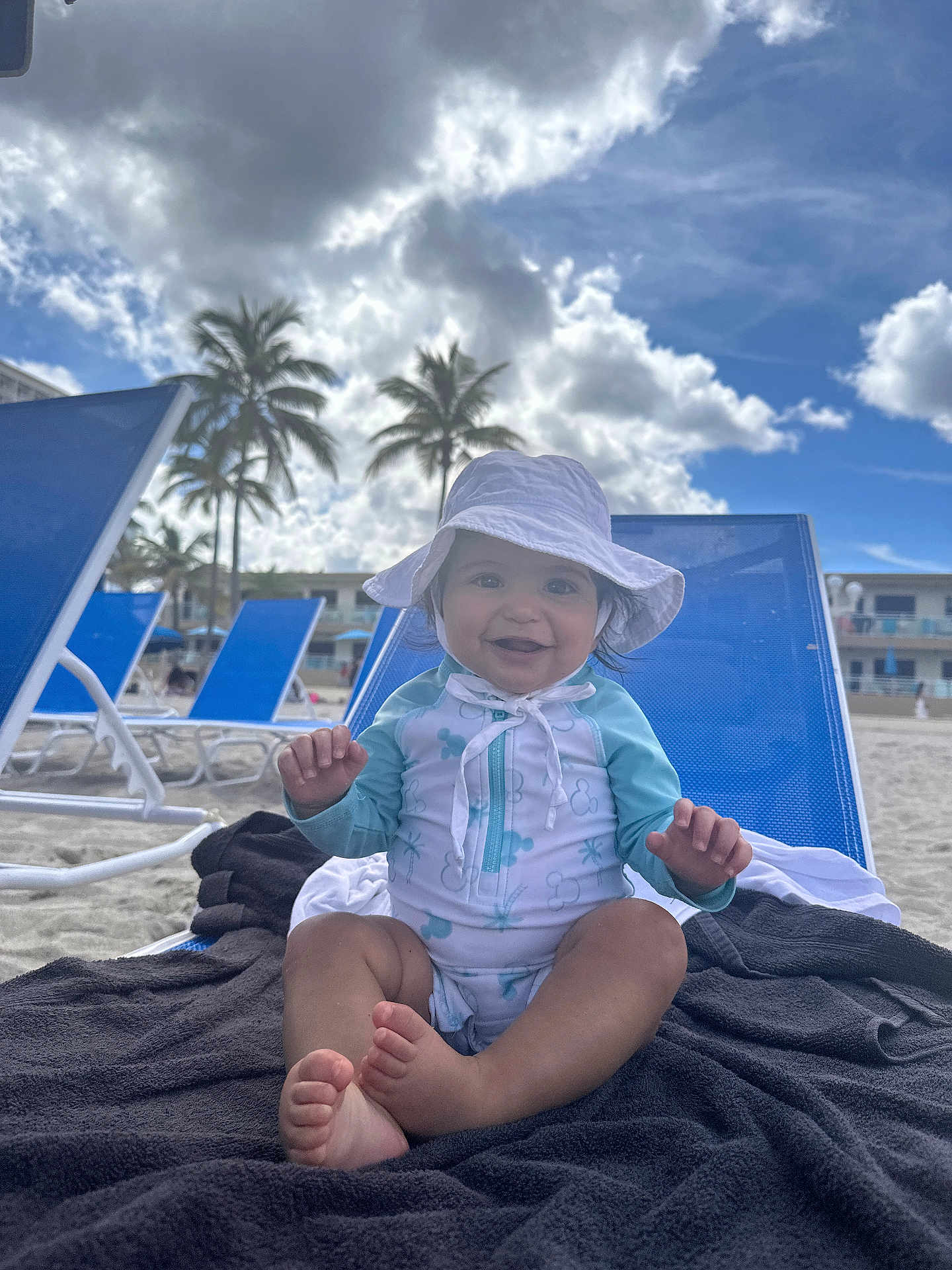 Renzo joined the competition — help win amazing prizes! baby, child, beach, sun_hat, swimsuit, towel, beach_chair, sand, palm_trees, sky, clouds, smiling, happy, outdoor, vacation, summer, sunny, person, foot, hand