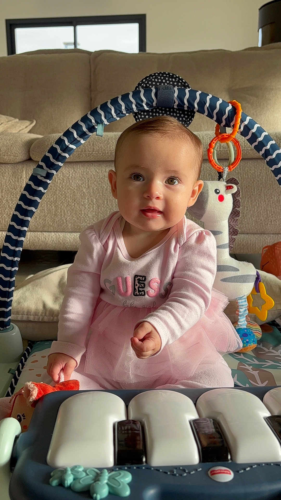 Liva a rejoint le concours — aidez-le/la à gagner de superbes lots ! baby, infant, child, play_mat, toy_piano, stuffed_toy, play_gym, teething_ring, pink_dress, tutu, sitting, smiling, big_eyes, living_room, couch, arch_toy, sunlight, adorable, portrait, hand