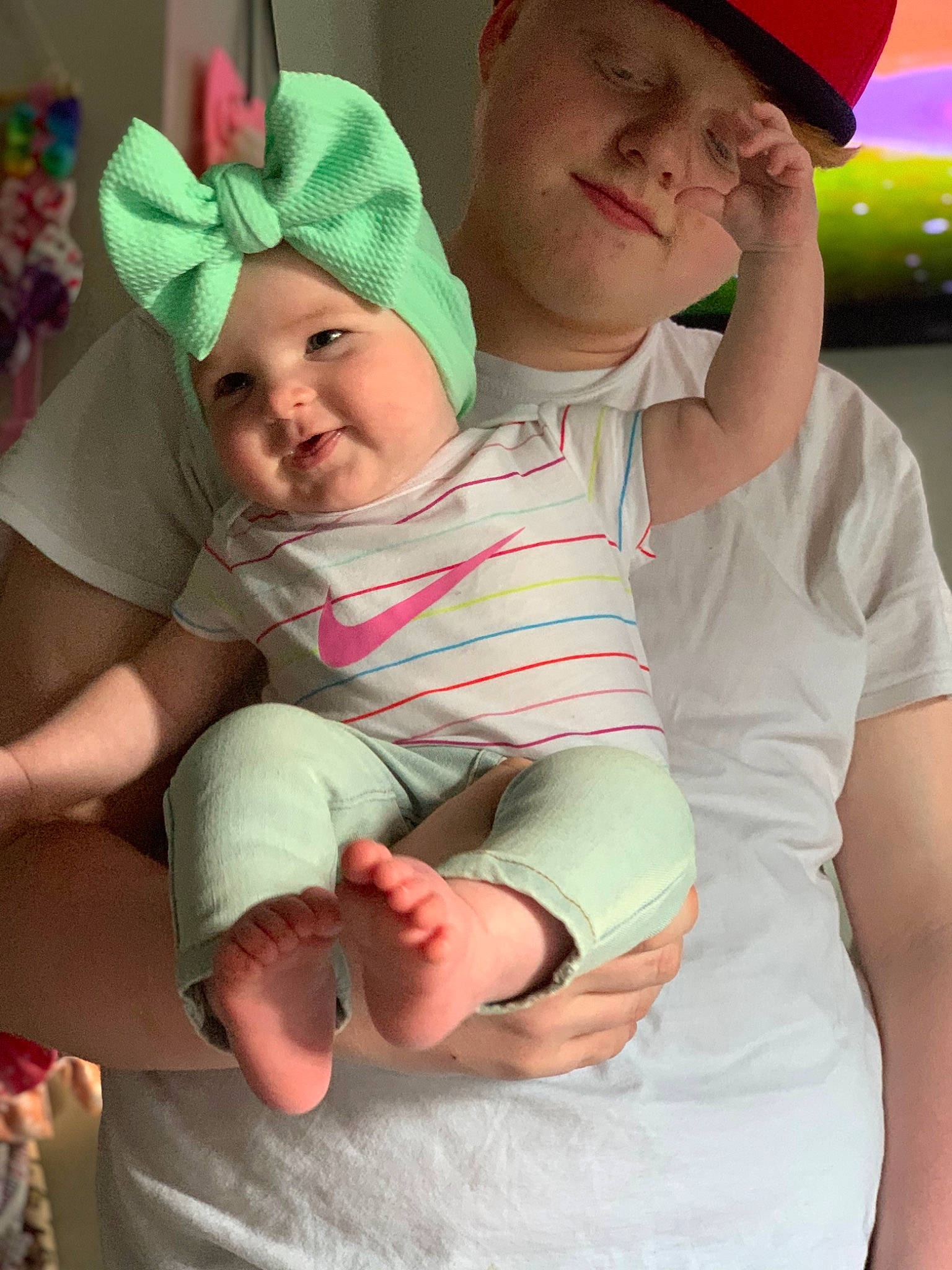 Delilah is registered to the contest to win money with this photo: baby, baseball_cap, cap, child, facial_expression, finger, fun, gesture, green, happy, hat, headgear, headwear, lap, mouth, organ, person, photograph, pink, smile