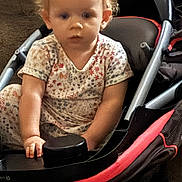 Lilly is registered to the contest to win money with this photo: baby, blonde_hair, child, closeup, dress, face, family, floral_clothing, indoor, infant, person, portrait, red_frame, serious, sitting, stroller, toddler, transportation, tray, wheel