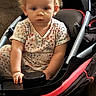 baby, blonde_hair, child, closeup, dress, face, family, floral_clothing, indoor, infant, person, portrait, red_frame, serious, sitting, stroller, toddler, transportation, tray, wheel