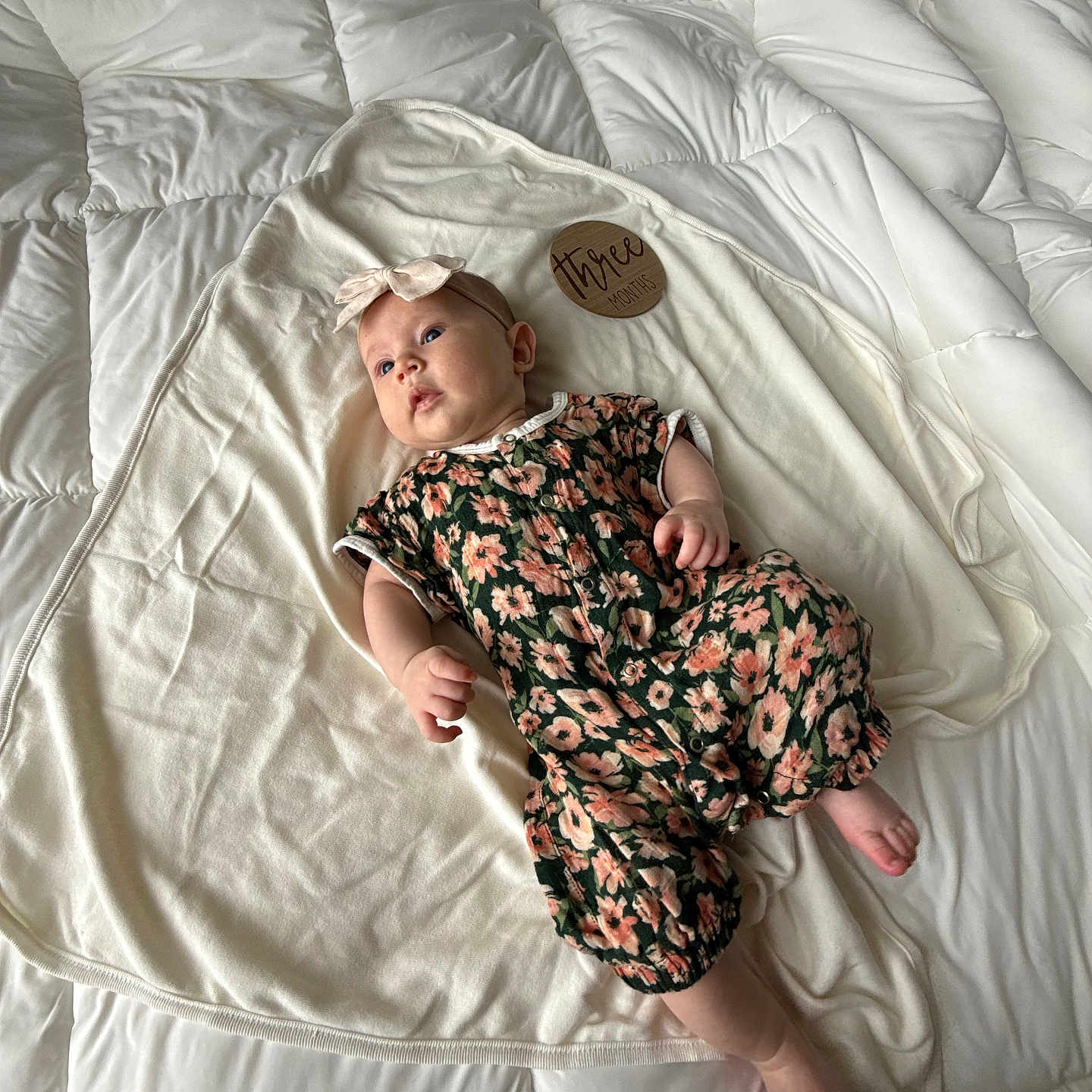 Magnolia is registered to the contest to win money with this photo: baby, infant, child, romper, headband, floral_pattern, blanket, bed, white, wooden_sign, three_months, lying_down, curious, soft_texture, comfort, indoor, person, cute, newborn, portrait