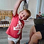 toddler, child, red_shirt, diaper, tongue_out, playful, indoor, crib, floor, legs, sitting, couch, tattoo, footwear, wooden_furniture, home, person, blur, light, casual