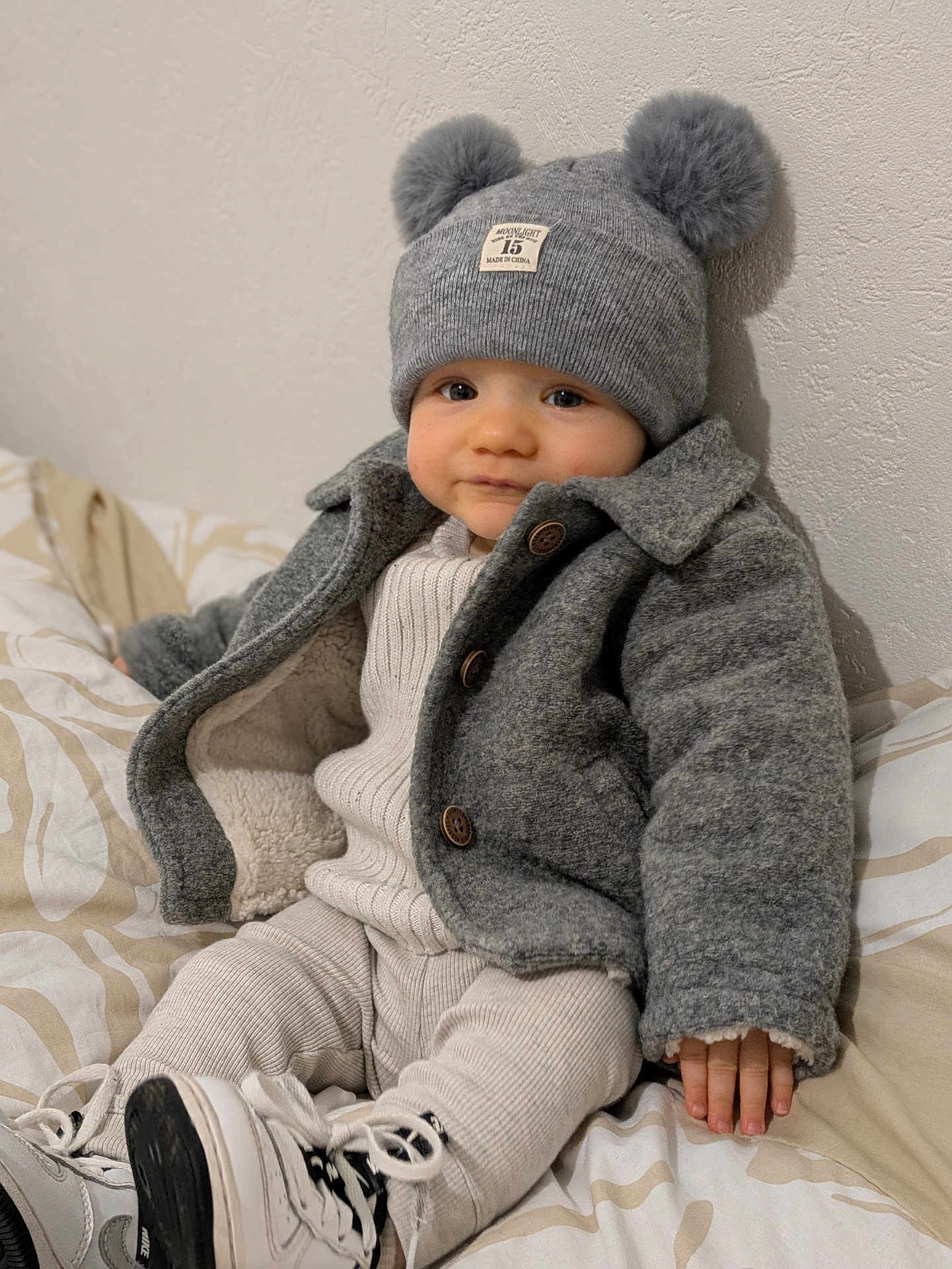 Malone participe au concours pour gagner de l'argent avec cette photo : baby, child, hat, pom_poms, coat, grey_clothing, sitting, bedspread, white_shoes, sneakers, indoor, curious_expression, warm_clothing, knitwear, buttons, patterned_bedspread, infant, cute, portrait, cozy