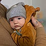 baby, child, knitted_hat, bear_costume, pacifier, adult_hand, warm_clothing, indoor, portrait, cute, soft_texture, close_up, cozy, toddler, face, expression, holding, person, infant, winter
