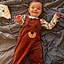 baby, child, overalls, brown_clothing, bear_design, blanket, gray_blanket, bed, smiling, reaching_hand, cute, indoor, infant, person, happy, cozy, clothing, long_sleeve_shirt, patterned_shirt, lying_down