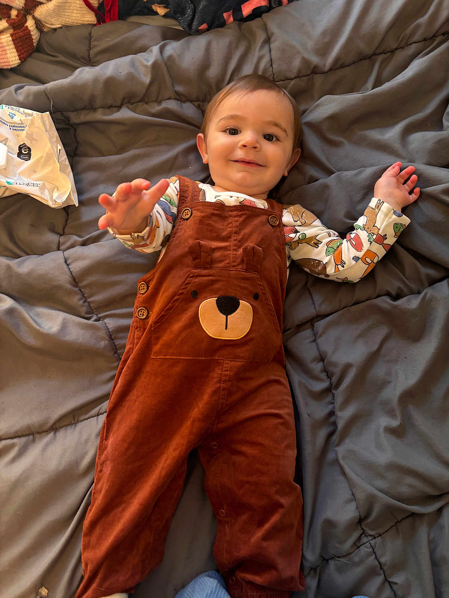 Colton joined the competition — help win amazing prizes! baby, child, overalls, brown_clothing, bear_design, blanket, gray_blanket, bed, smiling, reaching_hand, cute, indoor, infant, person, happy, cozy, clothing, long_sleeve_shirt, patterned_shirt, lying_down
