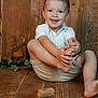 child, boy, smiling, happy, sitting, wooden_floor, wooden_wall, shorts, white_shirt, barefoot, pine_cones, toys, dominoes, greenery, indoor, portrait, cute, playful, natural_light, casual