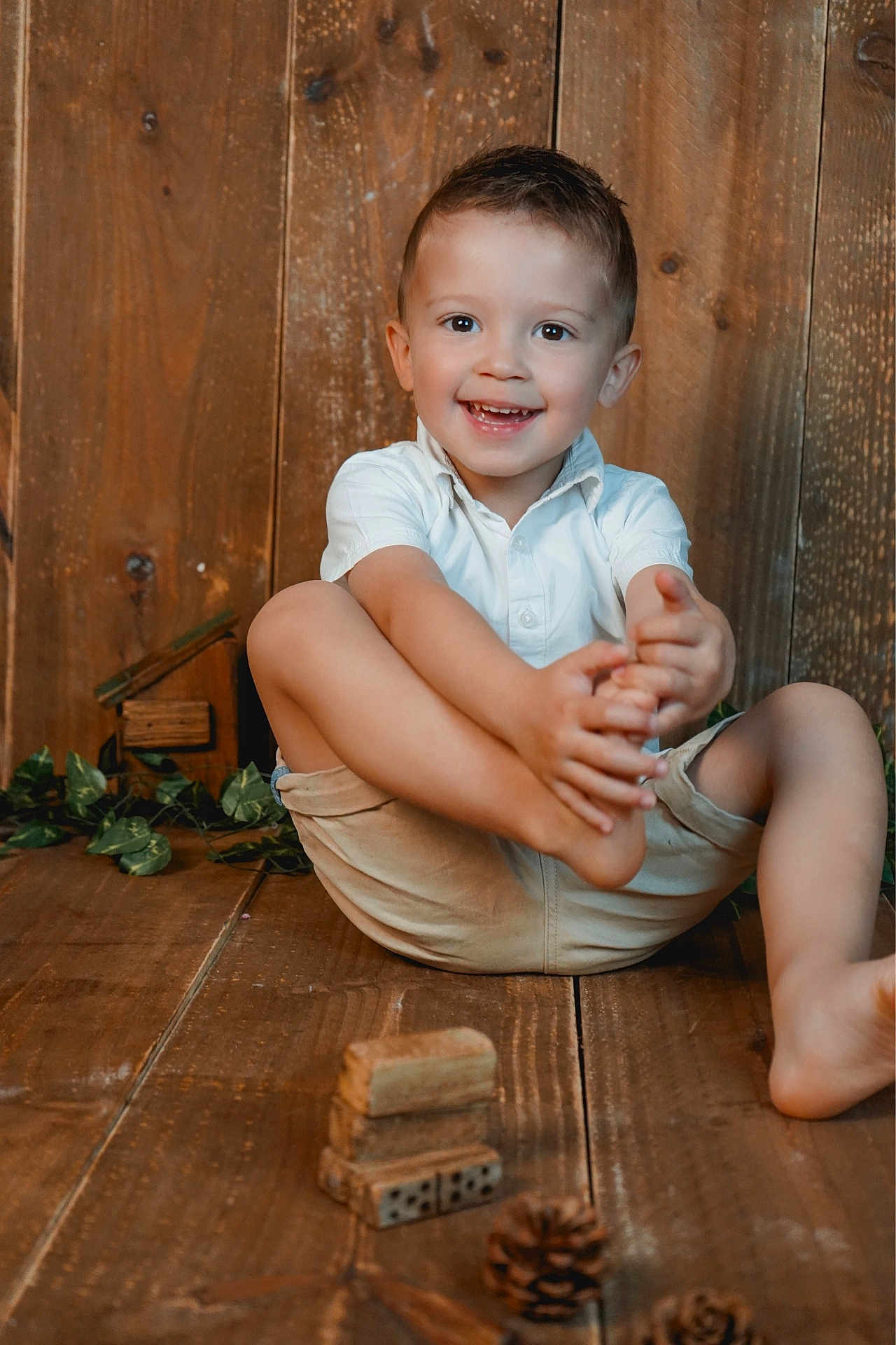 Côme participe au concours pour gagner de l'argent avec cette photo : child, boy, smiling, happy, sitting, wooden_floor, wooden_wall, shorts, white_shirt, barefoot, pine_cones, toys, dominoes, greenery, indoor, portrait, cute, playful, natural_light, casual