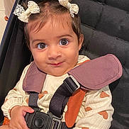 Samara is registered to the contest to win money with this photo: baby, child, stroller, safety_harness, bow, hair_accessory, cute, infant, clothing, patterned_outfit, white, orange, hand, face, eyes, portrait, indoor, seated, person, smile