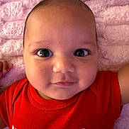 Dominic is registered to the contest to win money with this photo: baby, infant, close_up, face, eyes, red_shirt, text_on_shirt, blanket, pink, soft_texture, lying_down, child, cute, portrait, expression, skin, head, person, indoor, young