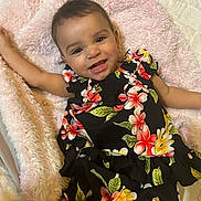 Kehlani is registered to the contest to win money with this photo: baby, child, smiling, floral_dress, blanket, soft_texture, cute, happy, lying_down, indoor, infant, person, portrait, toddler, colorful_clothing, skin, face, hand, head, cozy