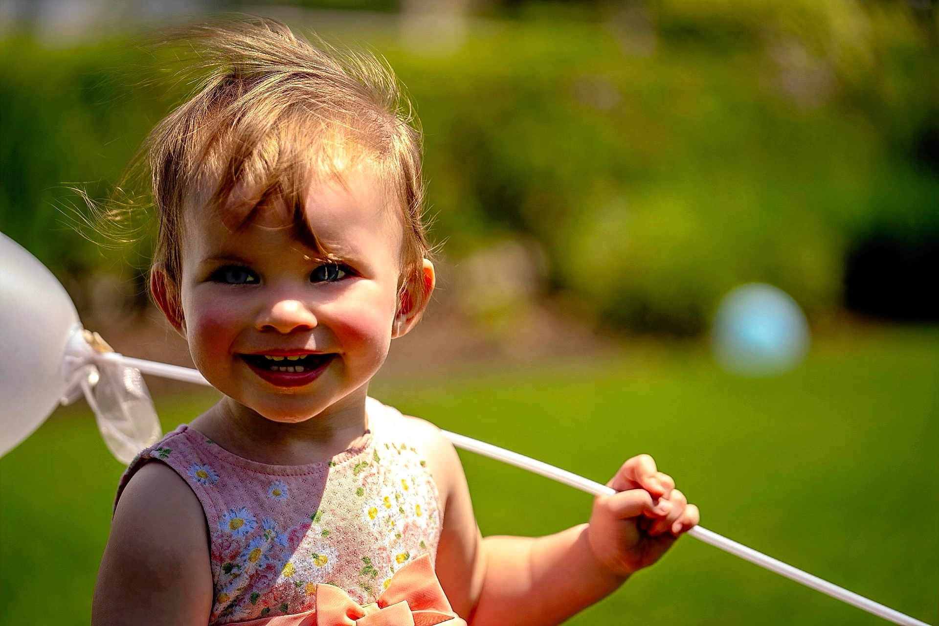 Willow is registered to the contest to win money with this photo: toddler, child, smiling, balloon, outdoor, greenery, sunlight, happy, face, portrait, dress, flower_pattern, rosy_cheeks, grass, nature, daylight, holding, cute, young_child, summer