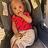 Cameron joined the competition — help win amazing prizes! baby, child, car_seat, puma, red_shirt, gray_pants, sitting, indoors, warning_label, curious_expression, hand_in_mouth, foot, infant, seatbelt, soft_padding, person, young_child, clothing, portrait, casual