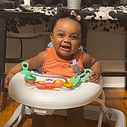 Milani is registered to the contest to win money with this photo: baby, child, toddler, smiling, happy, sitting, walker, activity_center, toy, playtime, indoor, home, speakers, audio_equipment, wood_floor, orange_clothing, portrait, infant, face, furniture