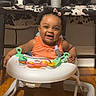 baby, child, toddler, smiling, happy, sitting, walker, activity_center, toy, playtime, indoor, home, speakers, audio_equipment, wood_floor, orange_clothing, portrait, infant, face, furniture