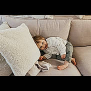 Leandre participe au concours pour gagner de l'argent avec cette photo : child, toddler, smile, couch, pillow, blanket, barefoot, shirt, pants, living_room, interior, cozy, soft_texture, portrait, playful, person, furniture, cushion, home, relaxed