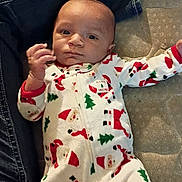 Marcello is registered to the contest to win money with this photo: baby, infant, child, face, onesie, pajamas, christmas_pattern, santa_print, lying_down, mattress, denim, jeans, leg, hand, eyes, cozy, indoor, portrait, cute, fabric