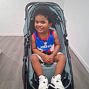 Justyce joined the competition — help win amazing prizes! child, toddler, smiling, stroller, portrait, indoor, home, wood_floor, wall, jersey, puerto_rico, sportswear, sneakers, white_shoes, necklace, bracelet, seated, curly_hair, happy, photography