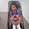 child, toddler, smiling, stroller, portrait, indoor, home, wood_floor, wall, jersey, puerto_rico, sportswear, sneakers, white_shoes, necklace, bracelet, seated, curly_hair, happy, photography