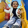 Cassie participe au concours pour gagner de l'argent avec cette photo : child, toddler, smiling, curly_hair, jacket, jeans, sneakers, playground, slide, outdoor, daylight, happy, person, casual_clothing, colorful, fun, reaching_hand, wood, metal, blue_sky
