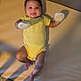 baby, infant, onesie, mittens, yellow_stripes, lying_down, smiling, happy, face, skin, legs, feet, bed, mattress, shadow, indoors, soft_light, curly_hair, child, person