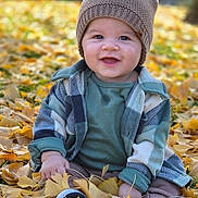 Bernardo is registered to the contest to win money with this photo: baby, child, smile, autumn, leaves, hat, plaid_jacket, outdoor, nature, seasonal, cute, sitting, fall, green_shirt, footwear, happy, young, portrait, sunlight, grass