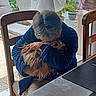 affection, blue_jacket, chair, comfort, cozy, dining_table, dog, elderly_person, embrace, glasses, home, hug, indoor, natural_light, pet, potted_plants, seated, tablecloth, watch, window