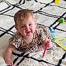 baby, carpet, child, crawling, cute, floor, floral_dress, home, indoor, infant, living_room, pattern, person, plush, red_object, rug, smiling, toy, yellow_card, young_child