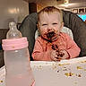 baby, child, chocolate, clothing, crumbs, cute, expression, face, feeding, food, hands, high_chair, indoor, infant, messy, milk_bottle, person, pink_bottle, smile, table