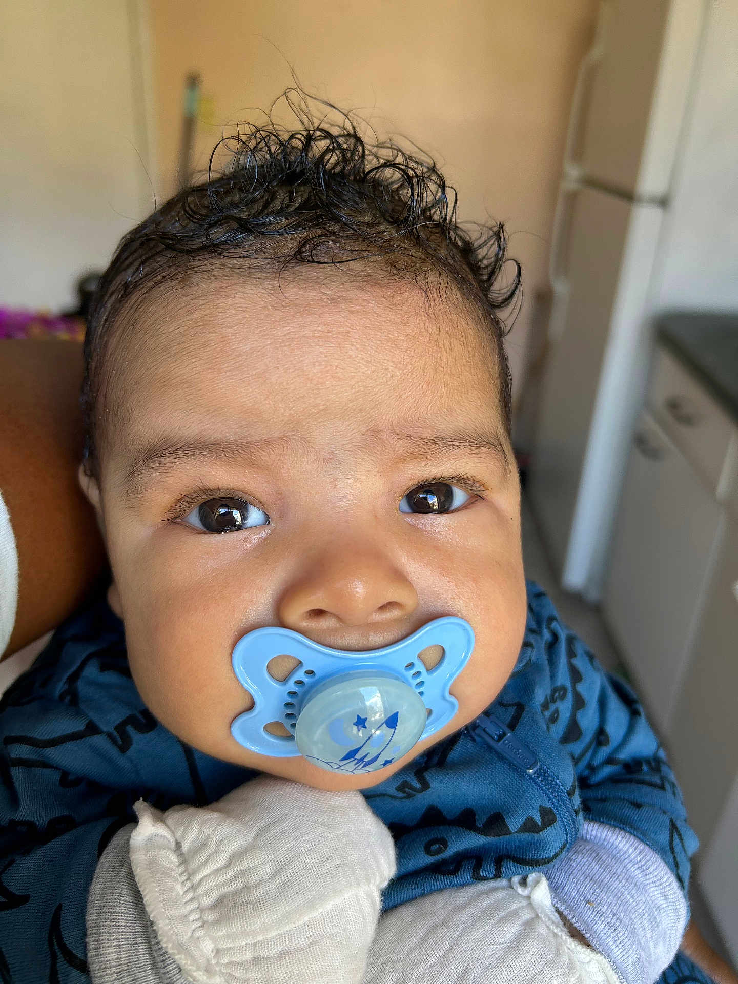 Angela joined the competition — help win amazing prizes! baby, infant, pacifier, blue_pacifier, close_up, face, eyes, curly_hair, onesie, mittens, hand, cheeks, portrait, indoor, kitchen_background, holding, cute, skin, expression, adorable