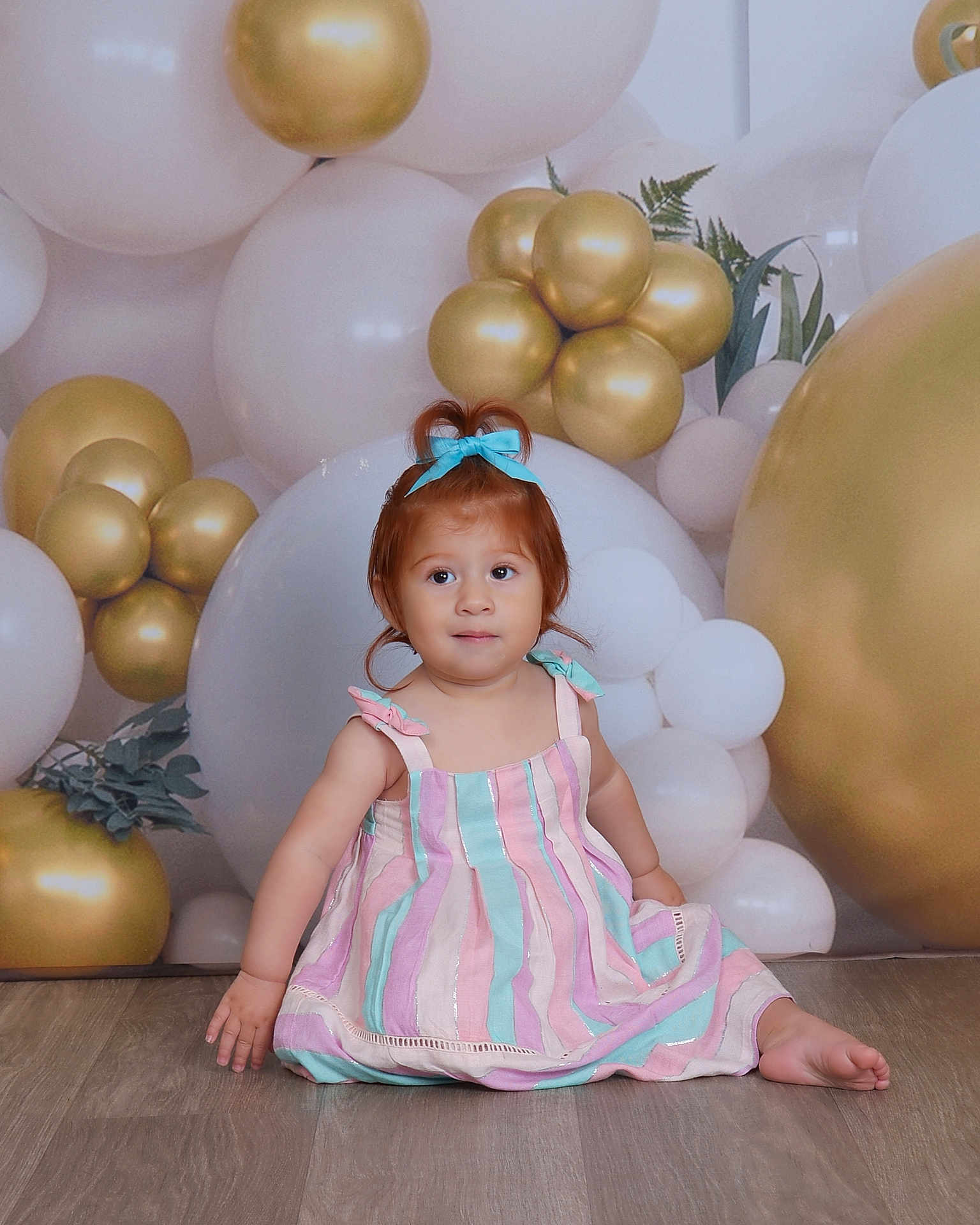 Andrea is registered to the contest to win money with this photo: toddler, child, balloons, gold, white, blue_bow, striped_dress, floor, indoor, portrait, cute, sitting, celebration, party, decor, young_child, smiling, person, flooring, happy