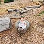 child, girl, outdoor, tree_stump, barefoot, dress, blue_bow, logs, leaves, nature, earth, curious, sitting, young, casual, daylight, ground, wood, play, relaxed
