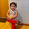 casual_clothing, child, crossed_legs, curly_hair, door, flooring, girl, happy, indoor, mat, pink_bows, polka_dot_shoes, portrait, posing, red_pants, small_stool, smiling, toddler, wall, wooden_floor