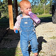 Ivy is registered to the contest to win money with this photo: toddler, child, denim_overalls, barefoot, bottle, toy_block, outdoor, sunlight, shadow, gravel, grass, tree, car, daylight, person, casual_clothing, nature, walking, curly_hair, curious
