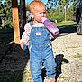toddler, child, denim_overalls, barefoot, bottle, toy_block, outdoor, sunlight, shadow, gravel, grass, tree, car, daylight, person, casual_clothing, nature, walking, curly_hair, curious