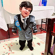 Veerjot is registered to the contest to win money with this photo: baby, blue_ribbon, carpet, chair, child, curly_hair, cute, floor, formal_wear, happy, indoor, person, play_mat, portrait, smiling, standing, suit, table, tie, toddler