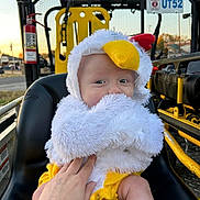 Caspian is registered to the contest to win money with this photo: baby, child, costume, chicken, fluffy, seat, hand, yellow, black, outdoor, vehicle, metal, sunset, face, person, cute, infant, portrait, clothing, expression
