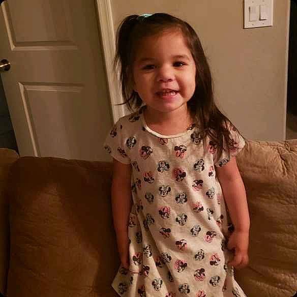 Emiliana is registered to the contest to win money with this photo: arm, brown_hair, cheek, child, dress, face, head, joy, long_hair, neck, nose, pattern, person, shoulder, sitting, skin, sleeve, smile, toddler