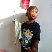 Moses joined the competition — help win amazing prizes! child, toddler, standing, stitch_tshirt, holding_cloth, blanket, bed, wall, sunlight, shadow, bracelet, curious_expression, portrait, indoors, sticker_on_wall, fabric, cloth, young_child, looking_up, single_person