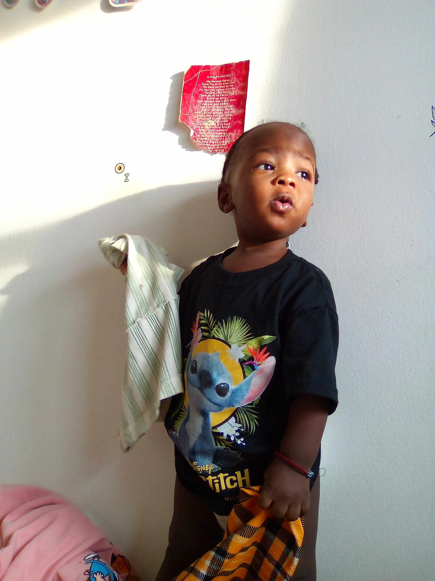 Moses joined the competition — help win amazing prizes! child, toddler, standing, stitch_tshirt, holding_cloth, blanket, bed, wall, sunlight, shadow, bracelet, curious_expression, portrait, indoors, sticker_on_wall, fabric, cloth, young_child, looking_up, single_person
